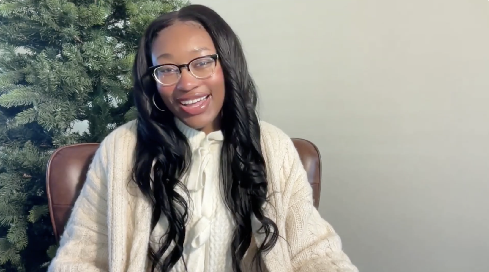A woman with long black hair, glasses, and hoop earrings, smiling while sitting in front of a holiday tree.