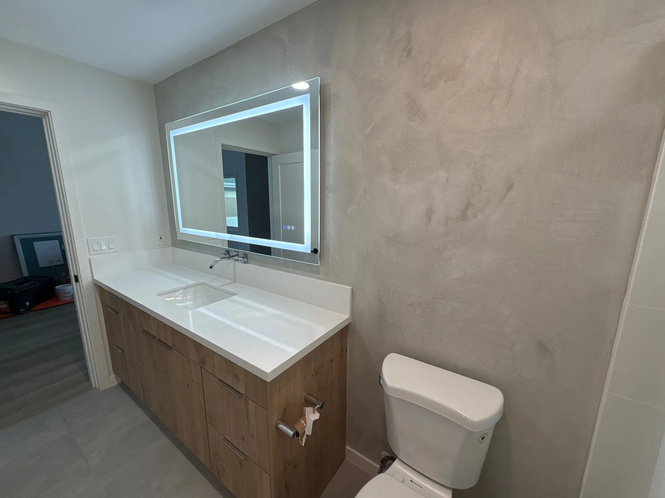 Modern bathroom with a wooden vanity, white countertop, microcement accent wall, a rectangular mirror with LED lighting, and a white toilet. Gray tiled floor and a textured gray wall.