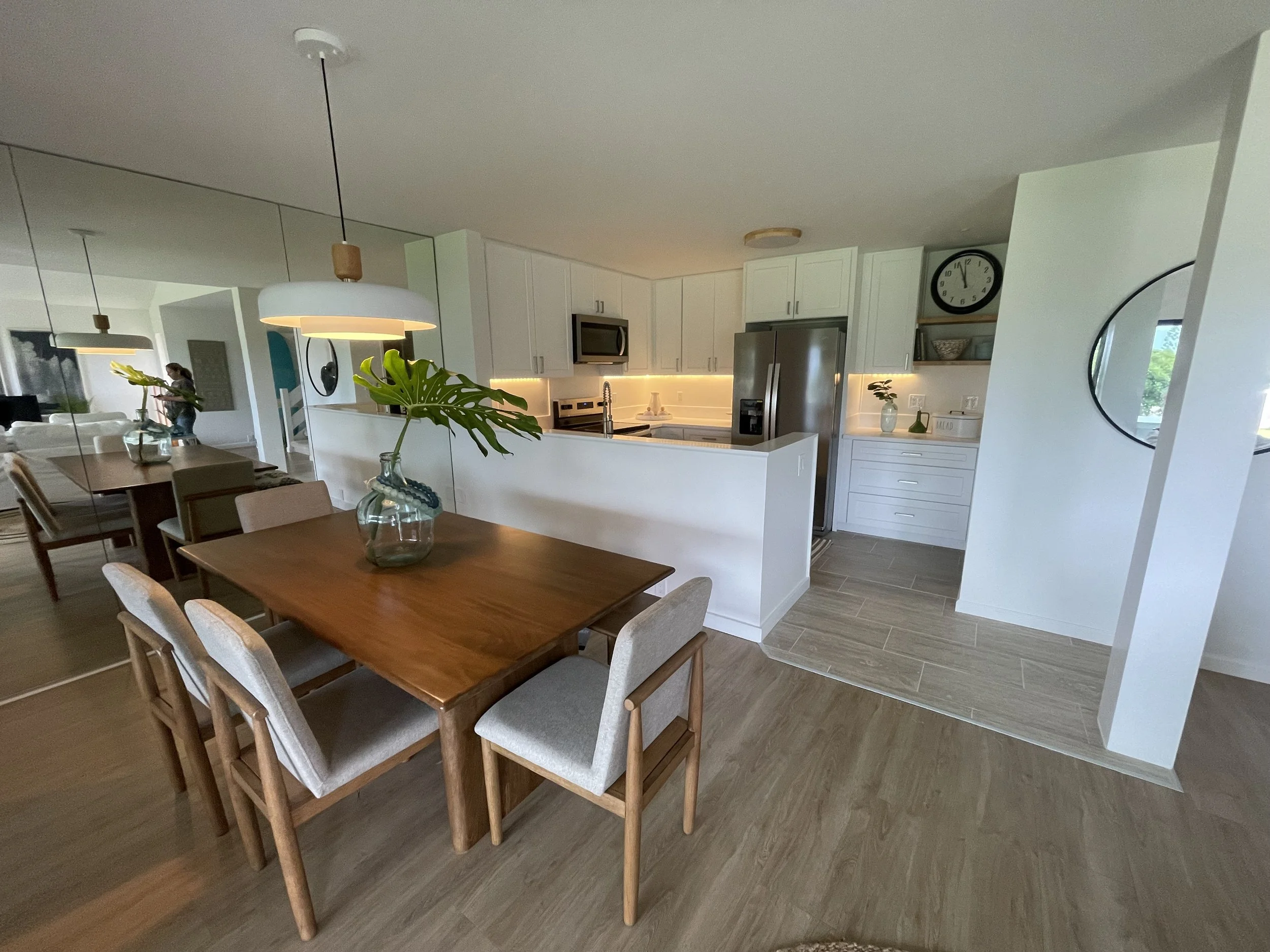 Open-concept kitchen and dining area with white cabinetry, stainless steel appliances, a wooden dining table with upholstered chairs, and a decorative vase with monstera plant leafs on the table. Mirror wall reflecting part of the room and natural li