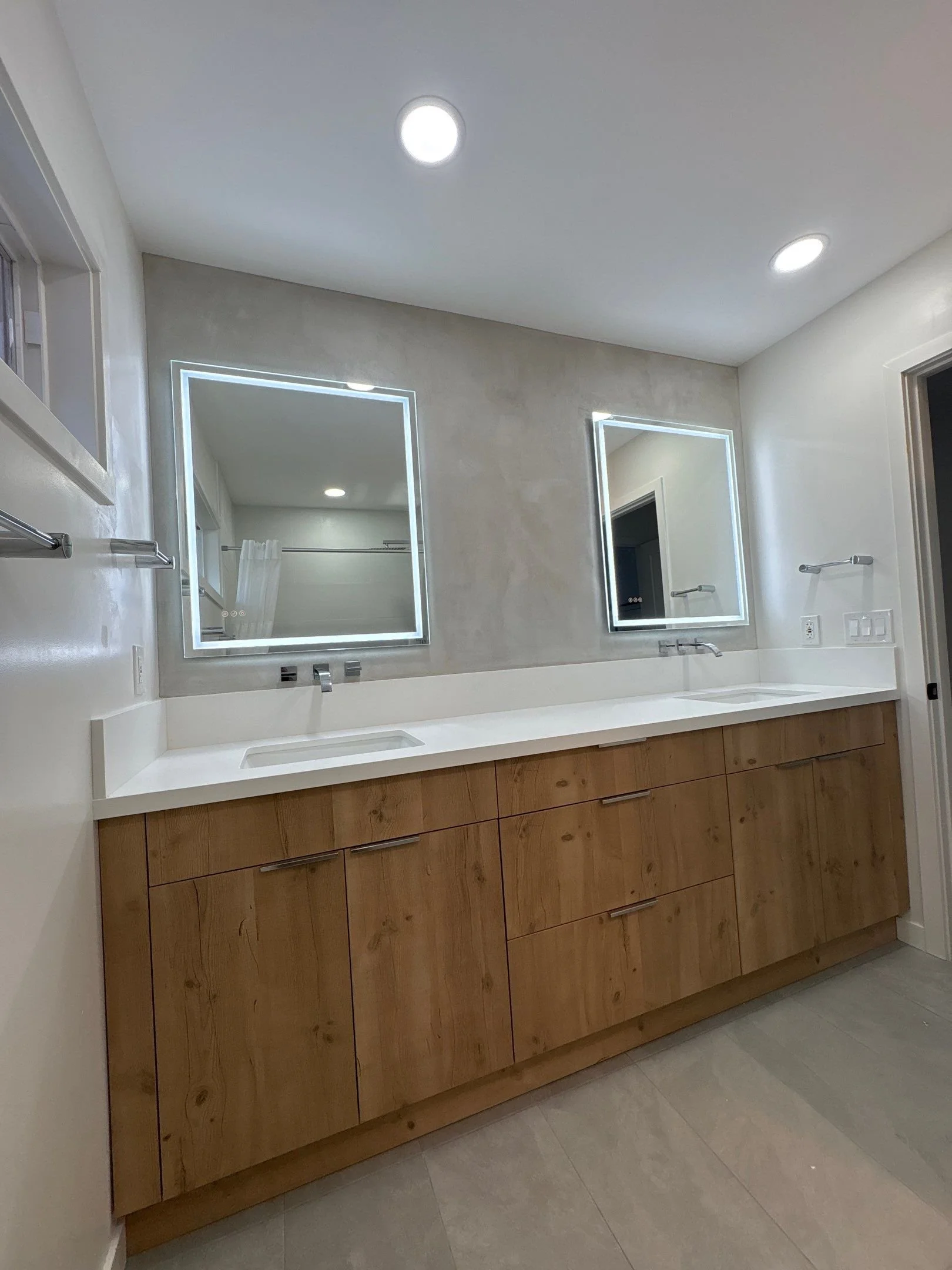 Modern bathroom with double vanity featuring a white countertop, microcement accent wall, wooden cabinets, two illuminated square mirrors, and recessed ceiling lights.