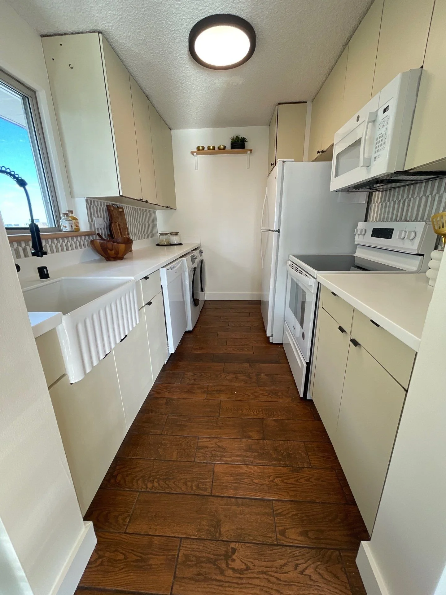 A narrow kitchen with white cabinets, wooden flooring, a window with a black sink faucet, appliances including a refrigerator, microwave, and stove, and a small shelf with plants and decorative items.