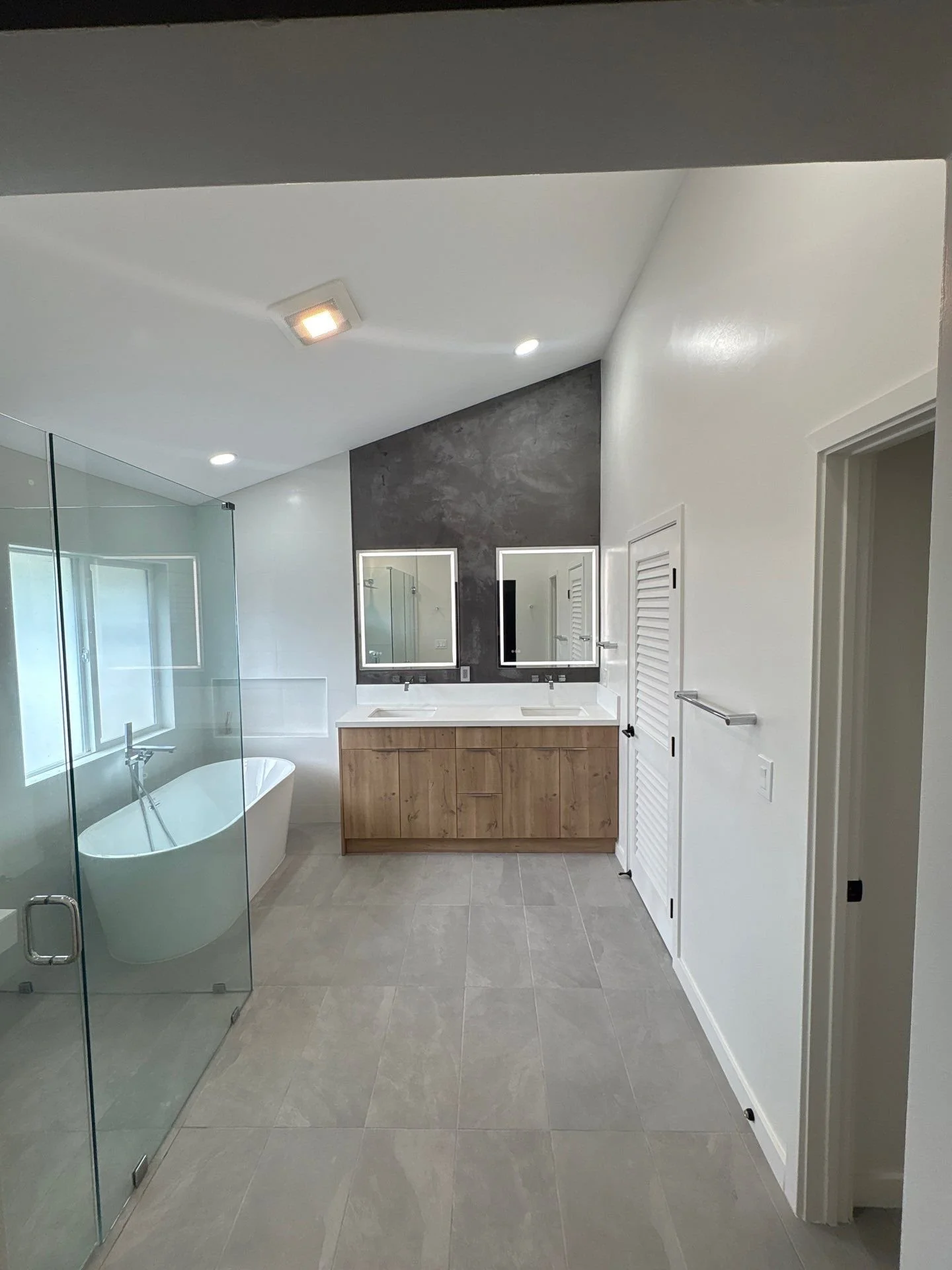 Modern bathroom with a standing glass shower, a freestanding bathtub, dual sinks with a wooden vanity, two mirrors, and a textured dark gray microcement accent wall.