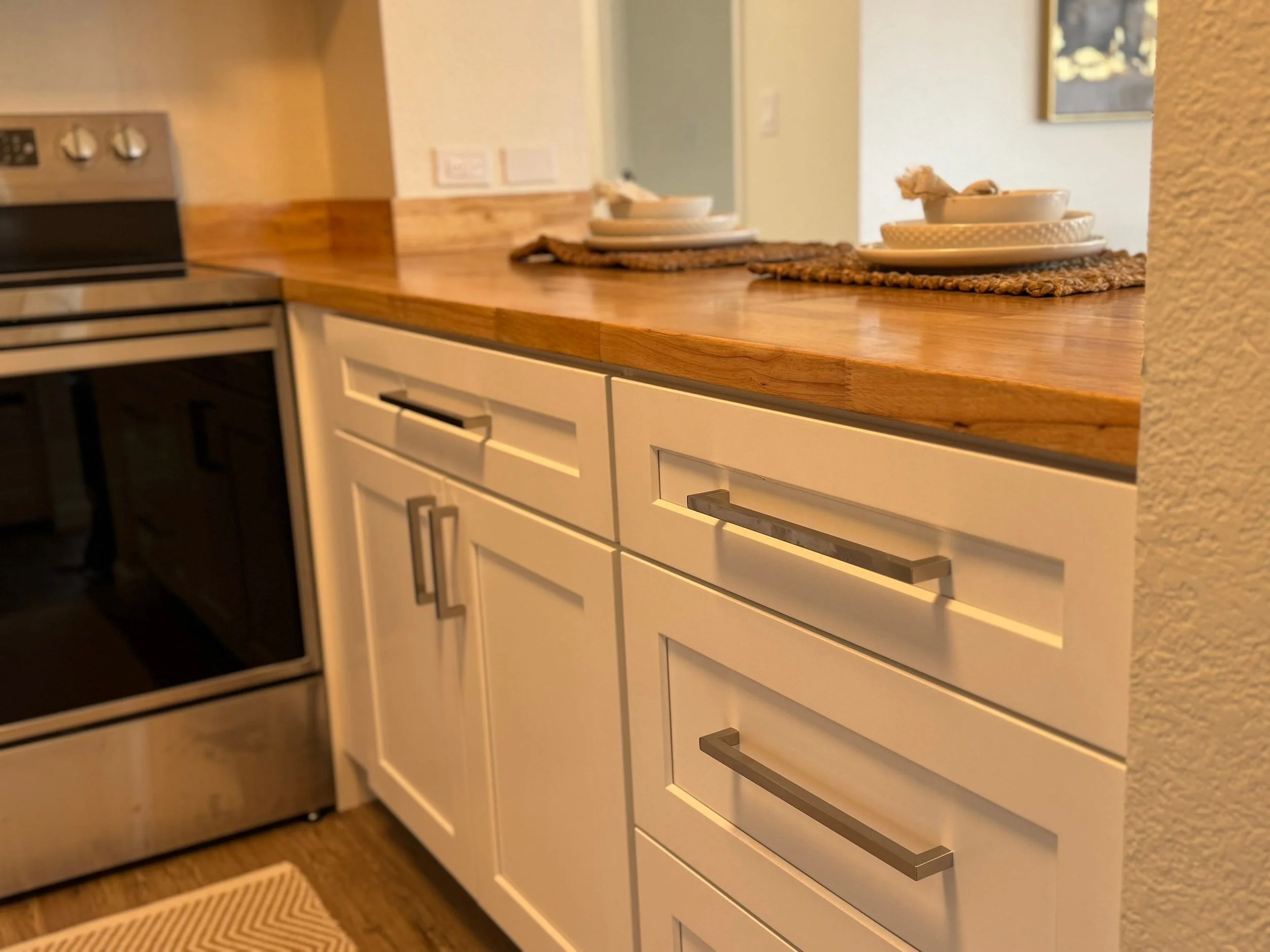 Close-up of a kitchen countertop with white cabinets, a stove on the left, and stacked white dishes on woven placemats on the right.