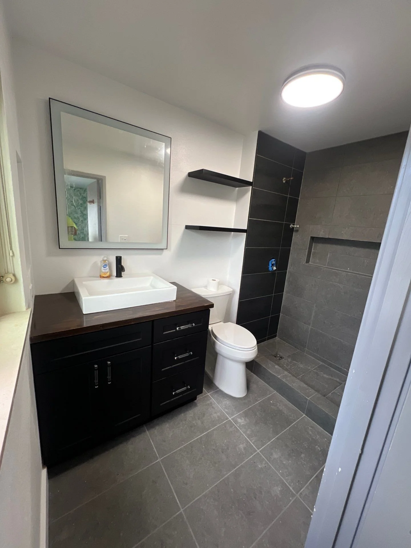 Modern bathroom with a black vanity, white sink, large mirror, toilet, and a walk-in shower with dark gray tiles and a small niche.
