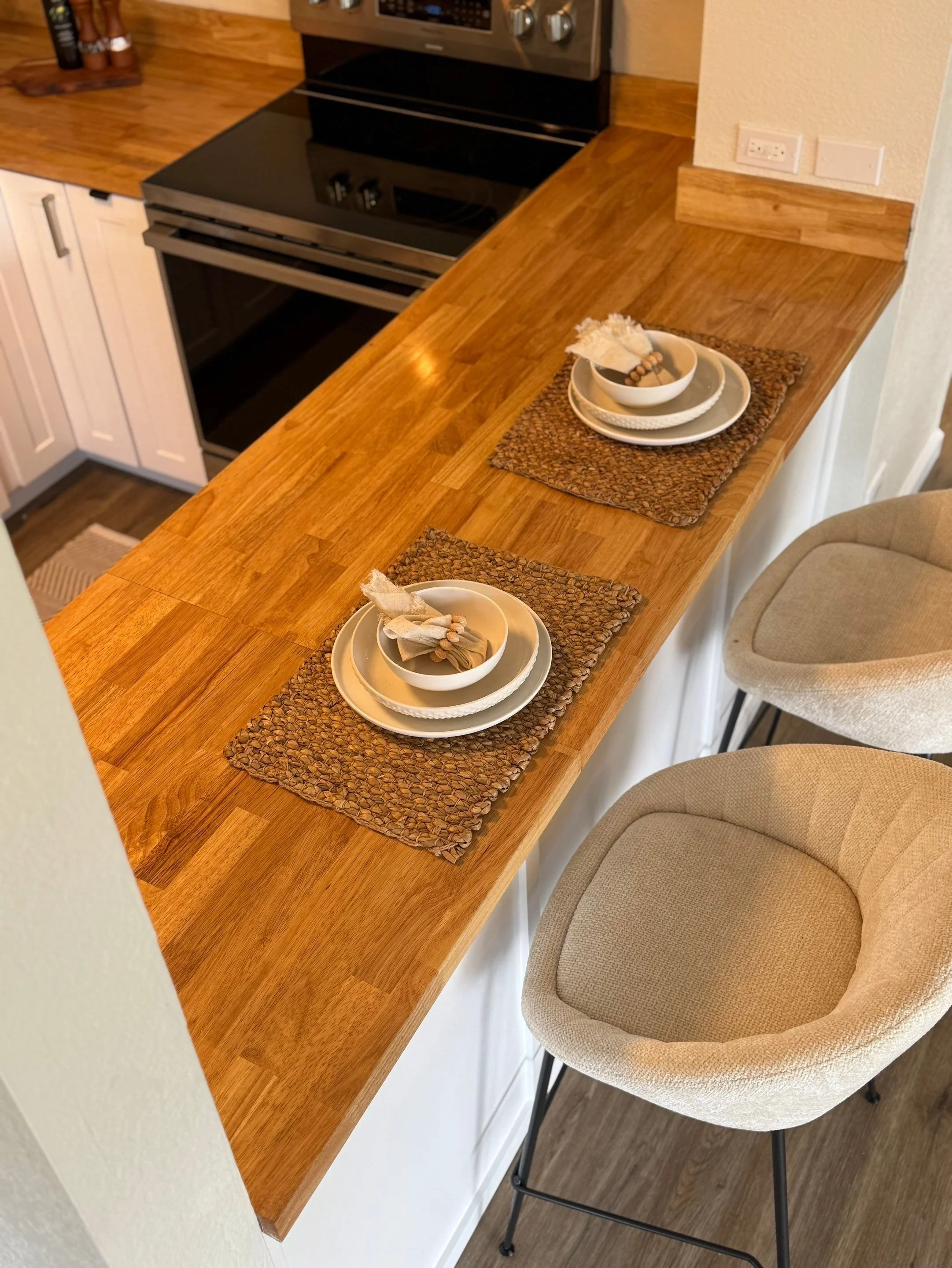 A wooden kitchen counter set with two place settings, each with a beige plate, bowl, napkin, and decorative items. Two beige chairs are positioned alongside the counter.