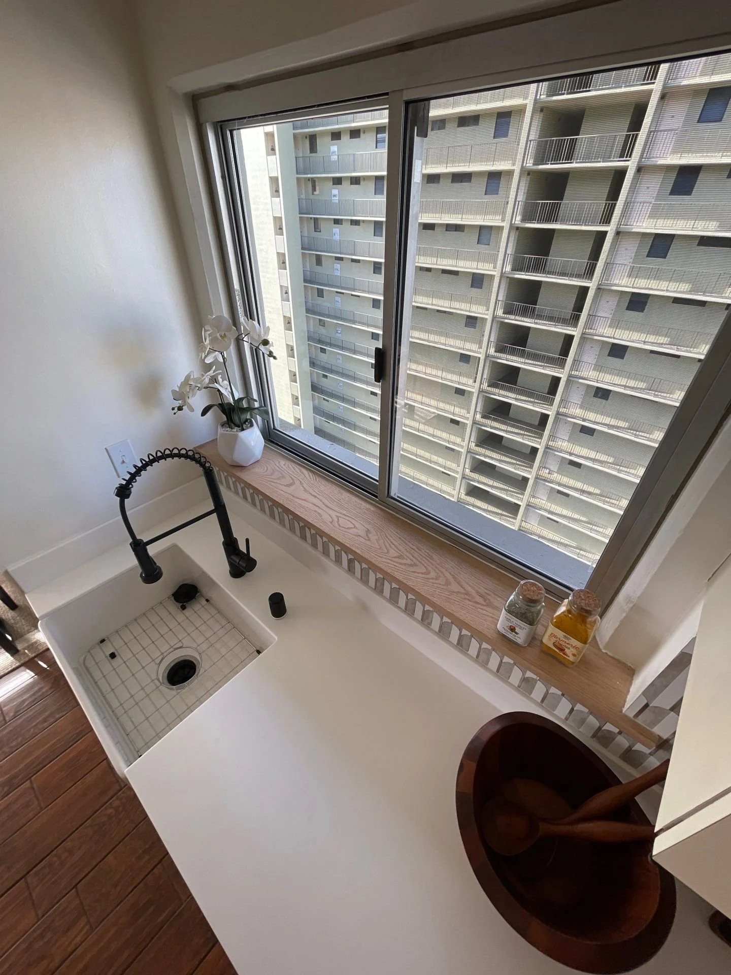 Kitchen sink with a black faucet, a potted white orchid plant on the window sill, and two jars near a window overlooking a high-rise apartment building.