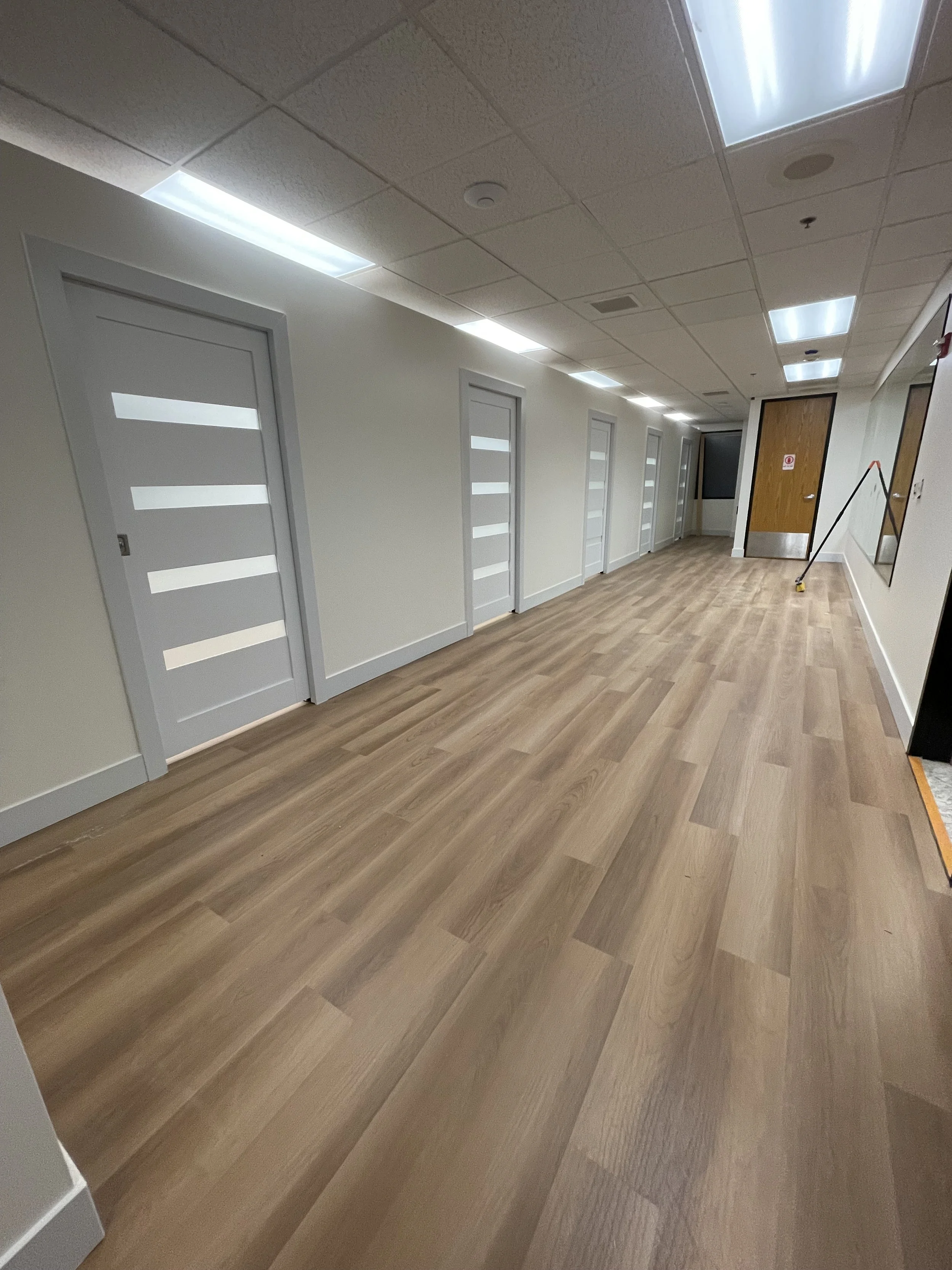 Empty hallway in a building with multiple closed doors on the left, a wood floor, bright ceiling lights, a mirror on the right wall, and a mop leaning against the wall.