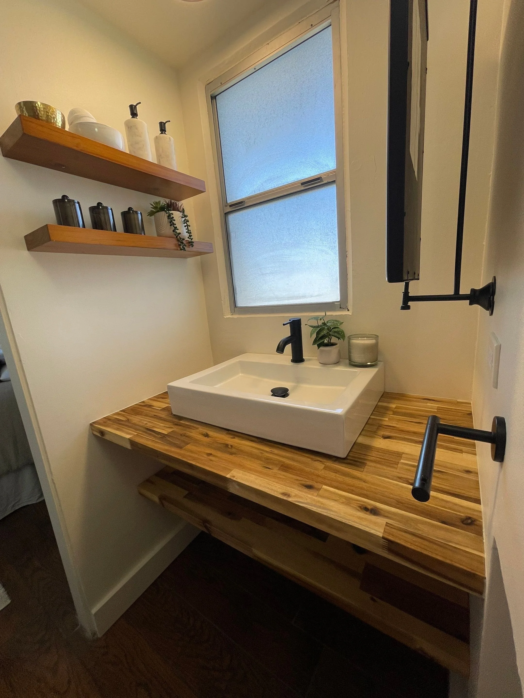 Bathroom vanity with a vessel sink, black faucet, wooden countertop, window, wall-mounted shelf with decor, and a small mirror.