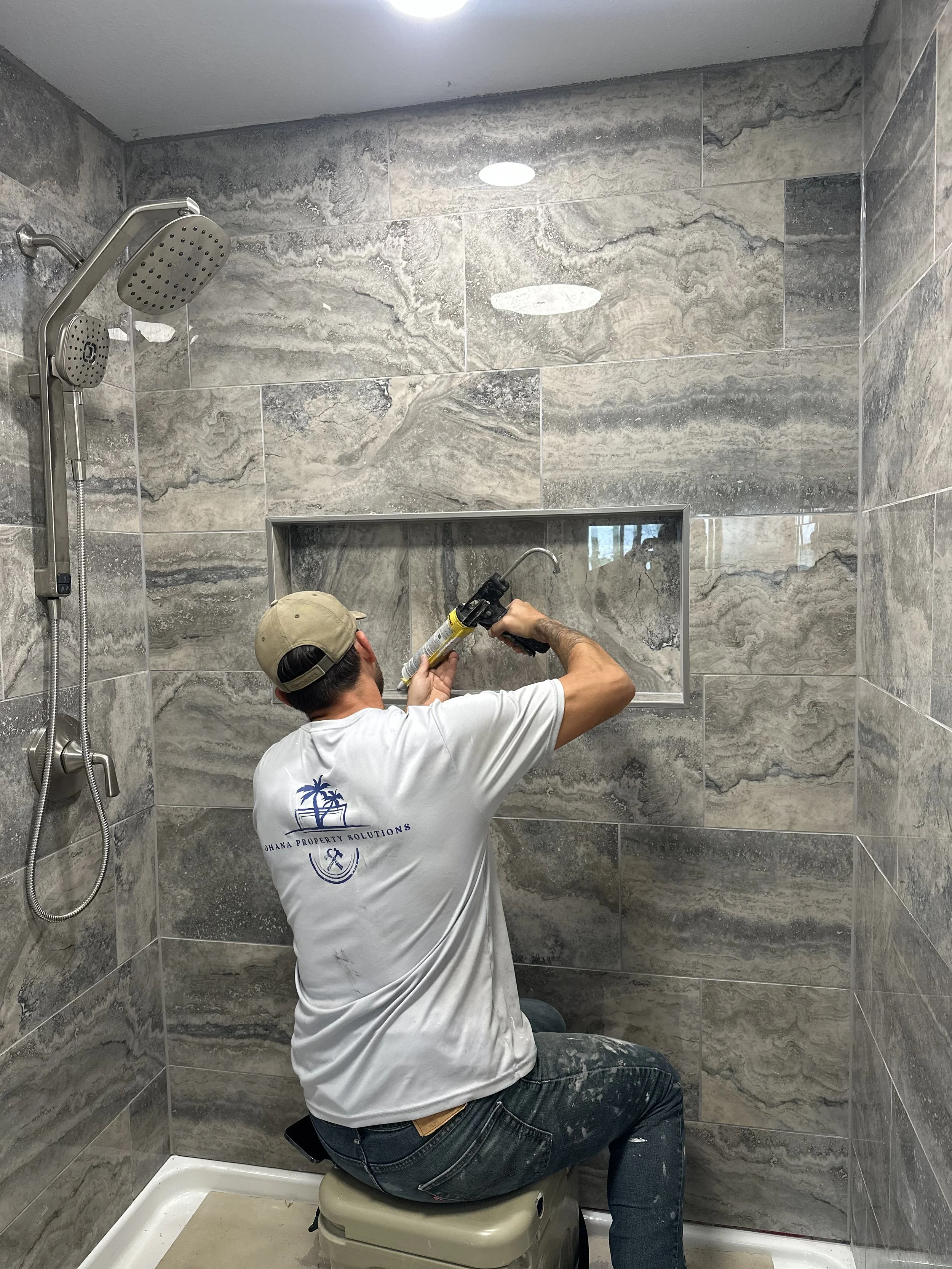 A man installing or repairing a shower with gray marble tiles, sitting on a toilet in a bathroom, wearing a beige cap, white T-shirt, and jeans, using a caulking gun.