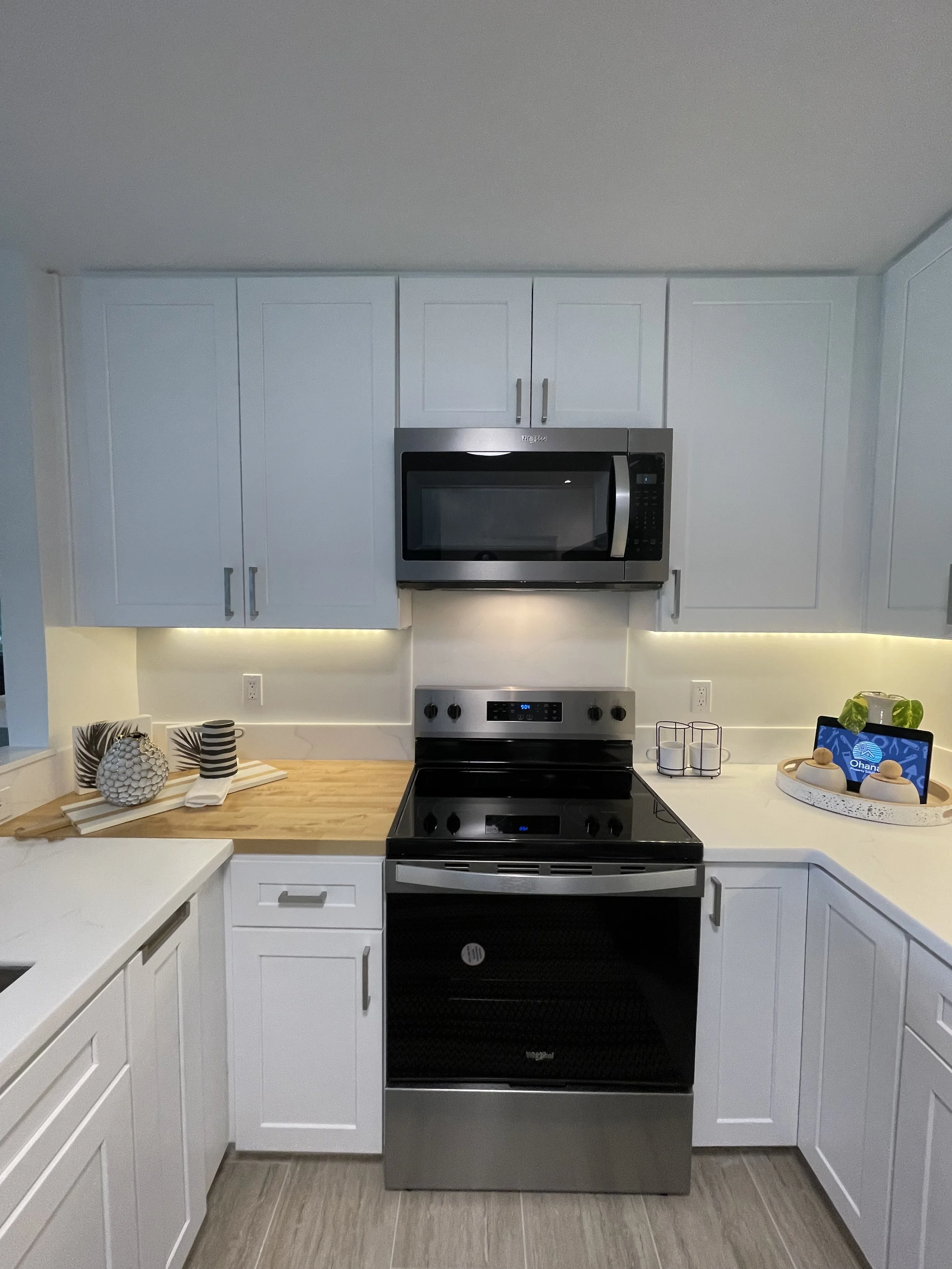 Modern kitchen with white cabinets, stainless steel microwave and stove, wooden and white countertops, decorative items, and a tablet on a tray.