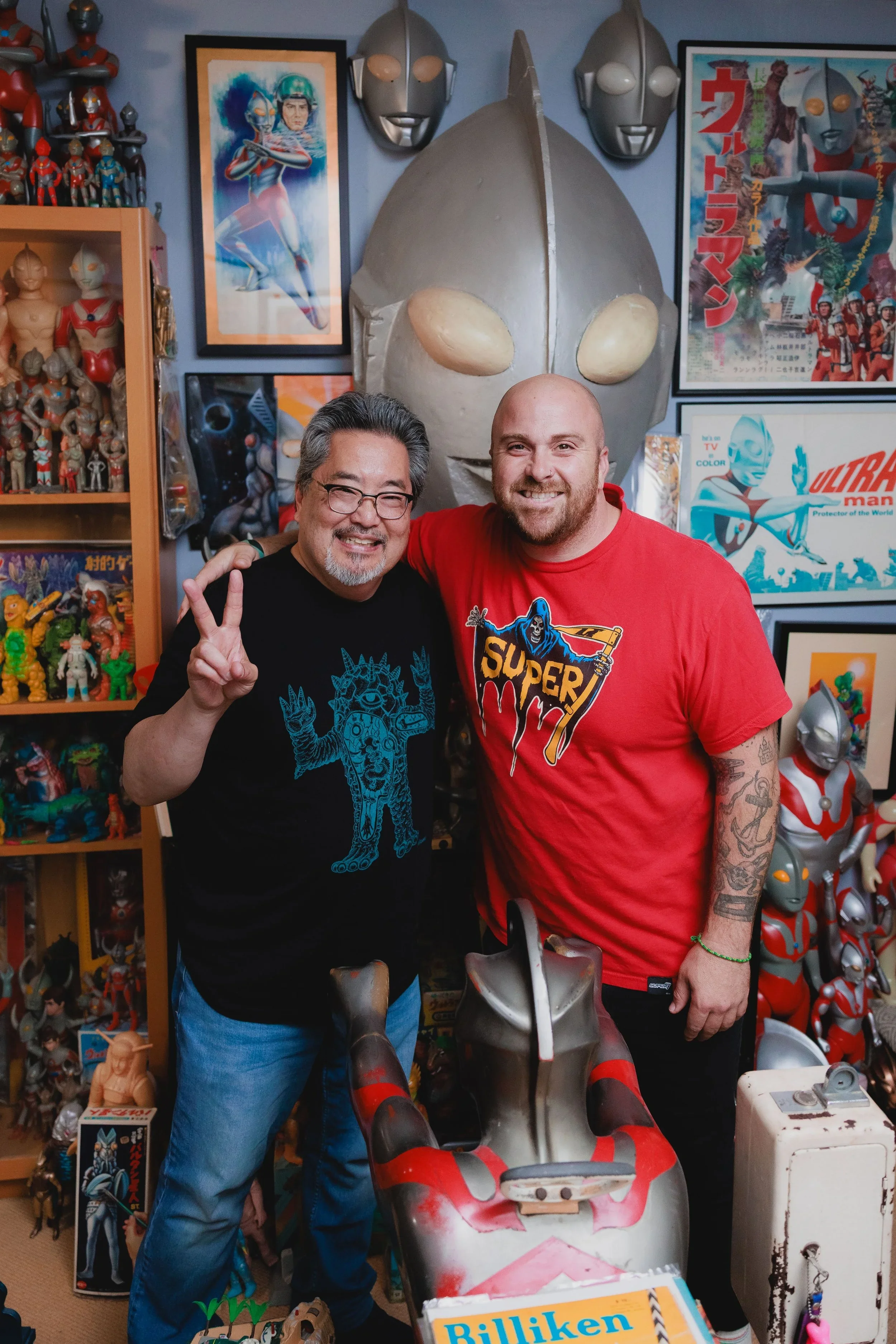 Two men in a room filled with vintage and collectible Ultraman and superhero memorabilia. One man is wearing glasses and a black shirt with a monster design, and the other man has a shaved head and a red shirt with a superhero logo. They are smiling and posing with an Ultraman figure in front of them. The room is decorated with Ultraman posters, masks, and action figures.