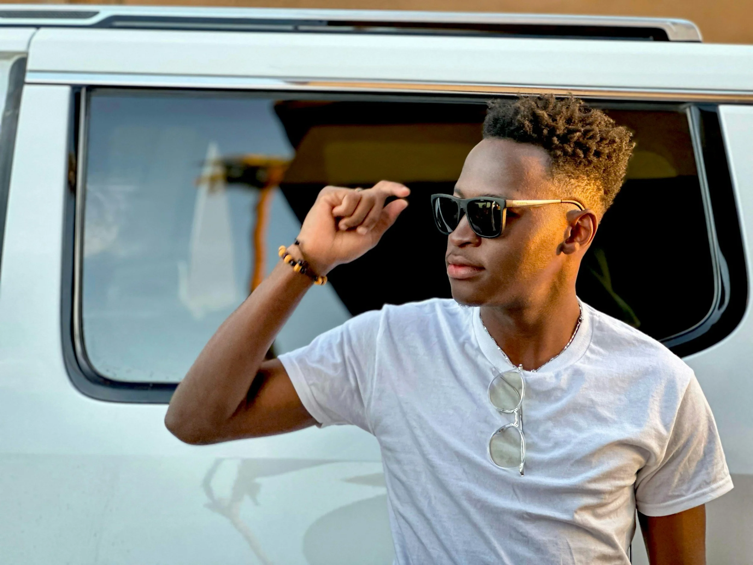 Young man with sunglasses and a white t-shirt standing outdoors near a vehicle, adjusting his sunglasses.