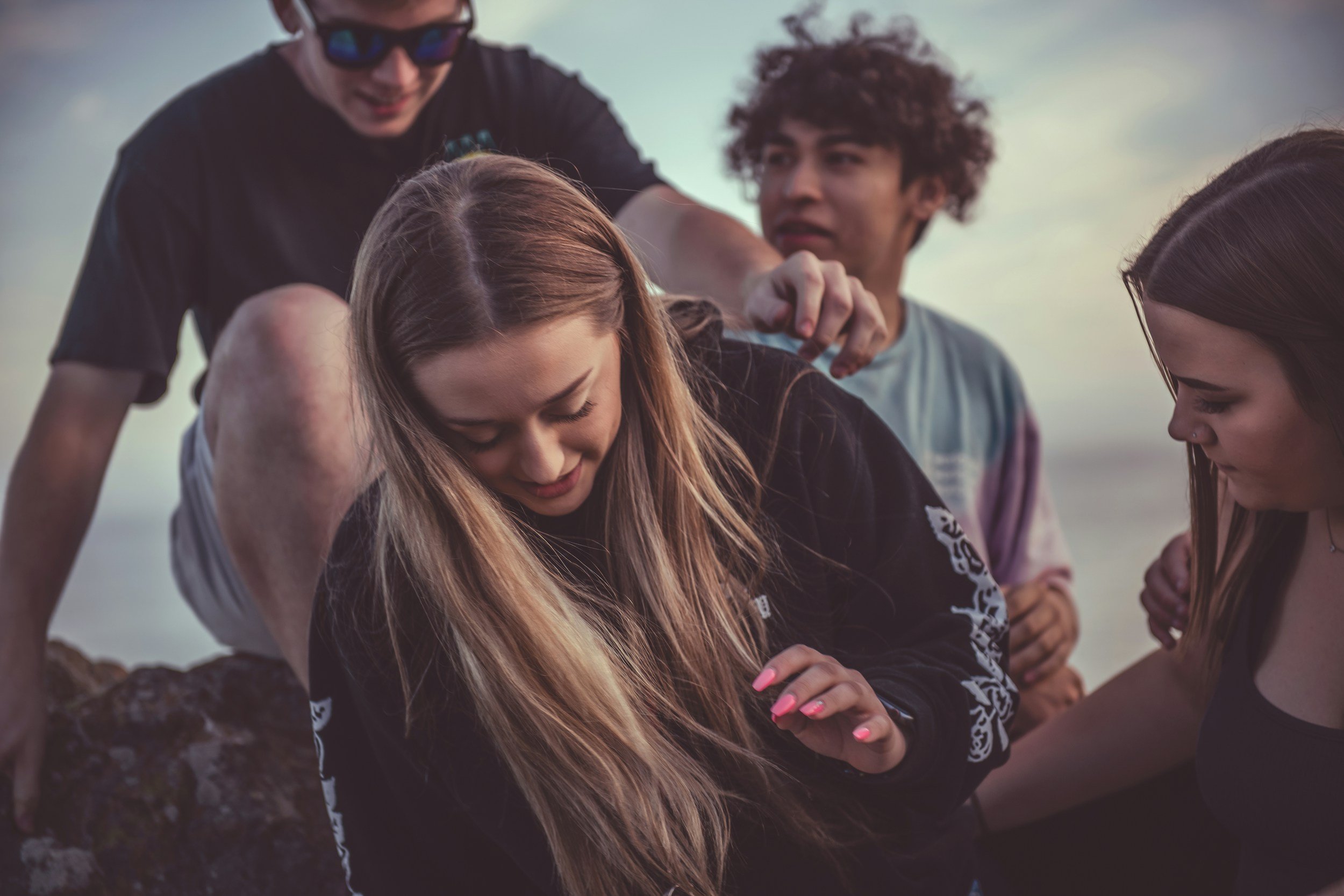 Group of four young people outdoors, one girl in black clothing smiling, with one boy helping her from behind, and another girl looking down at something beside her, during sunset or overcast sky.