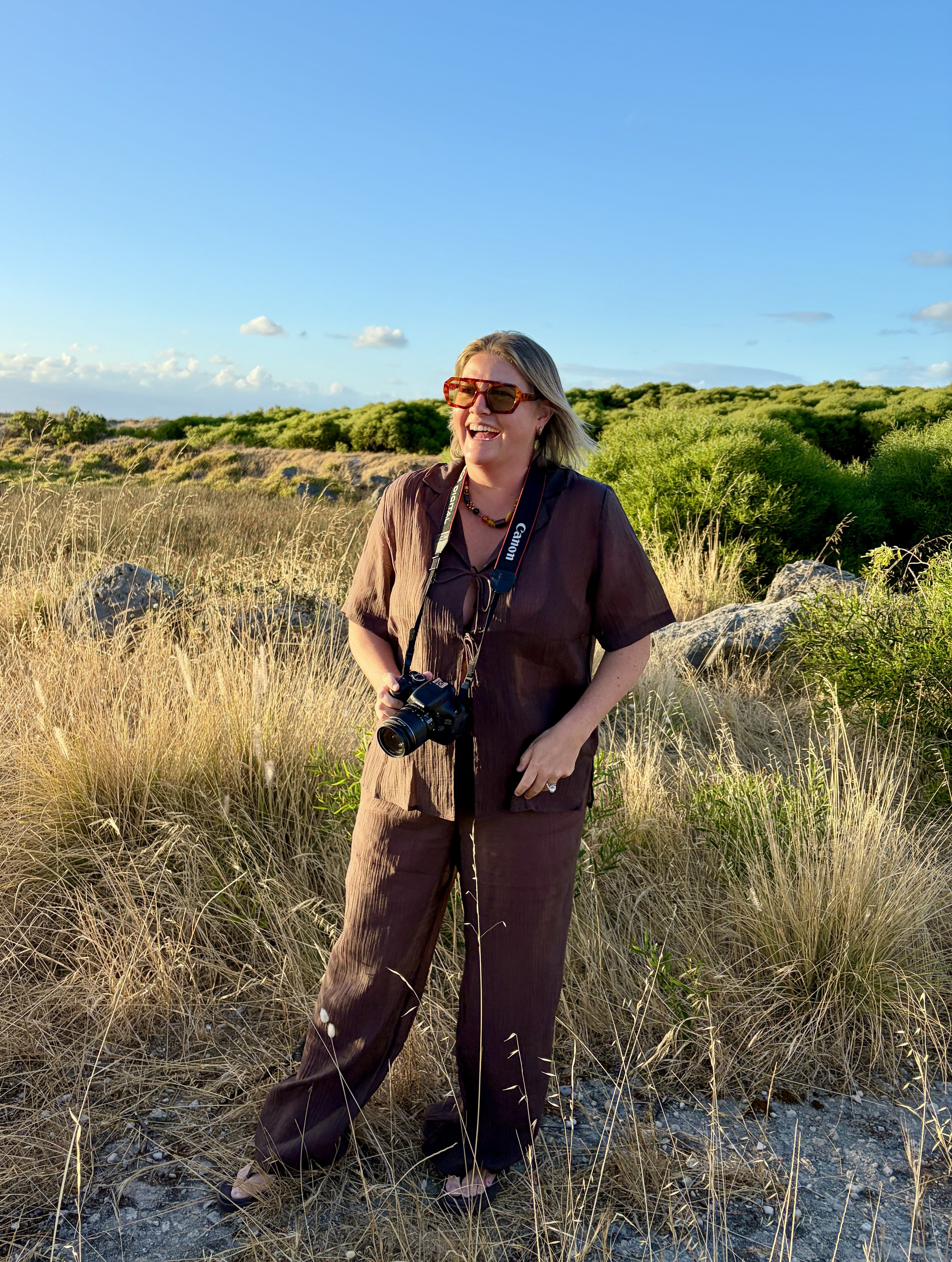 A woman standing outdoors in a grassy field, smiling, with a camera around her neck, wearing sunglasses, a dark brown outfit, and earrings, under a bright blue sky with some clouds.