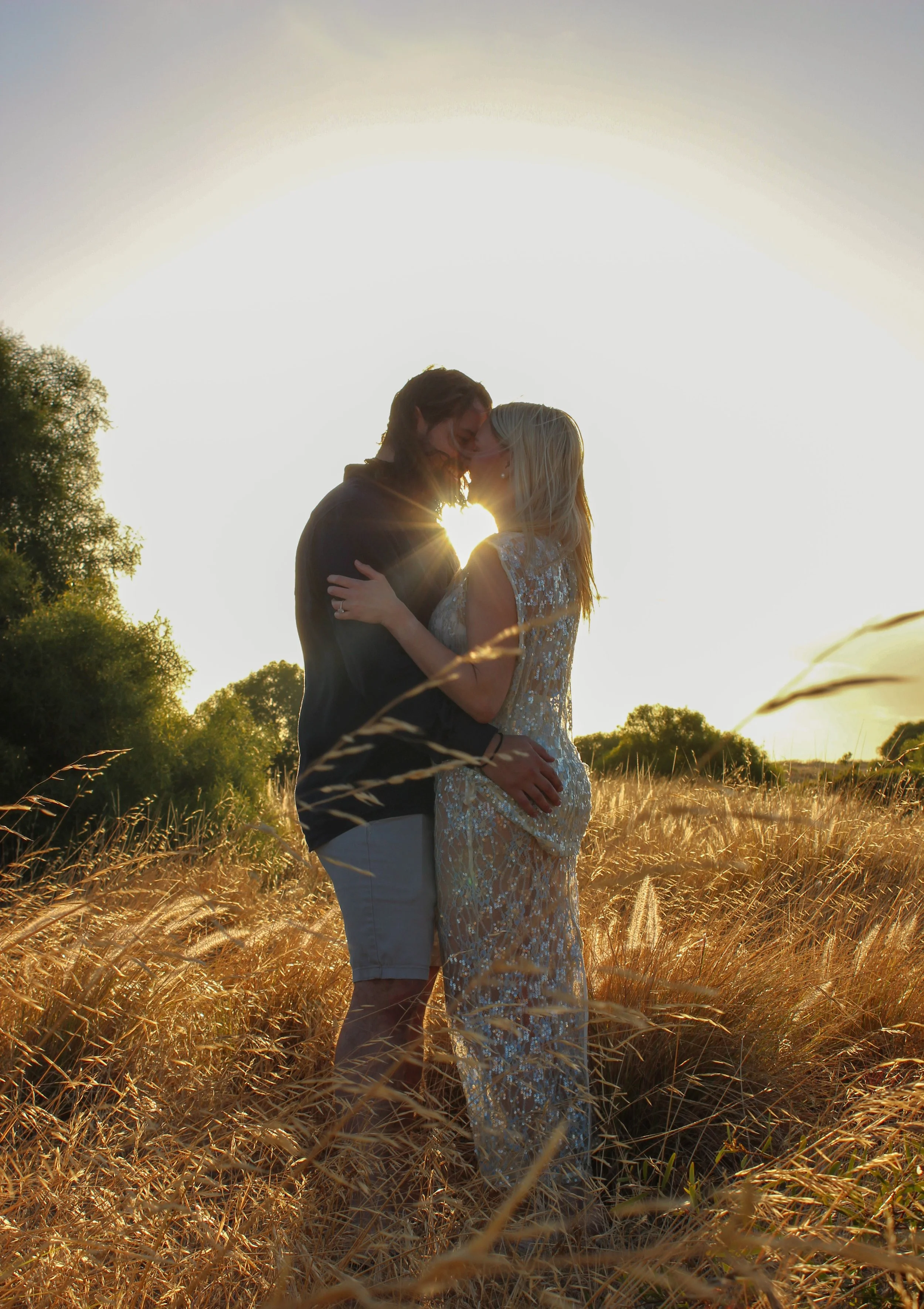 A couple standing close together in a field of tall grass at sunset, with their foreheads touching and the sun shining behind them.