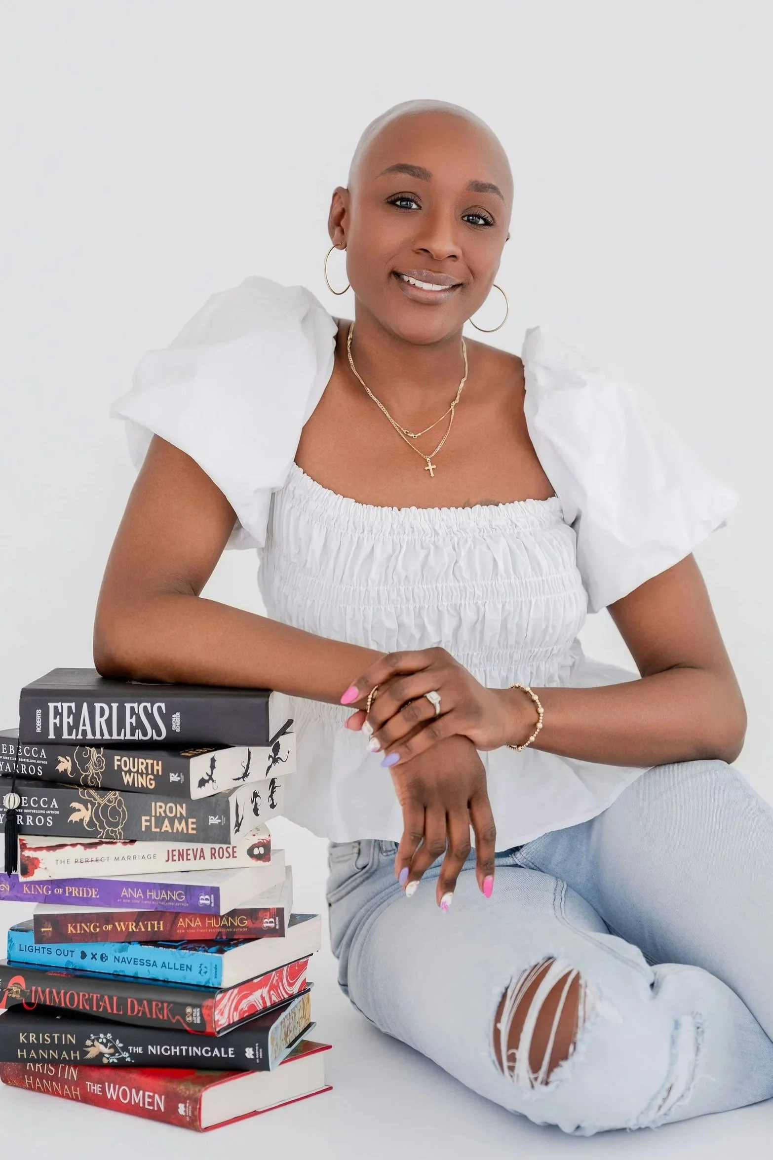 A woman with a shaved head, wearing a white puff-sleeve top and ripped jeans, sitting next to a stack of books and smiling at the camera.