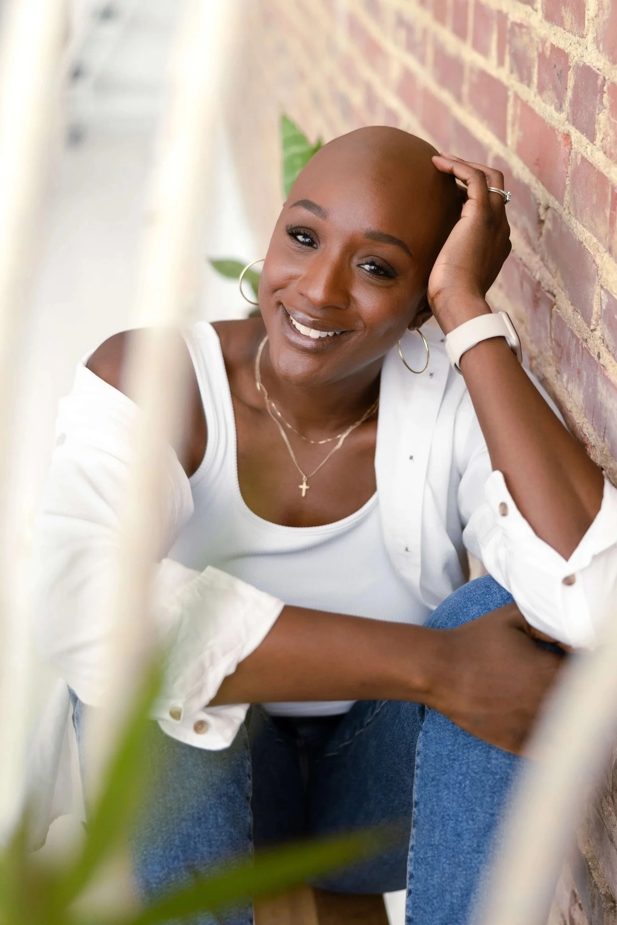 A woman with a shaved head sitting against a brick wall, smiling at the camera, wearing a white top, hoop earrings, a cross necklace, and a watch.