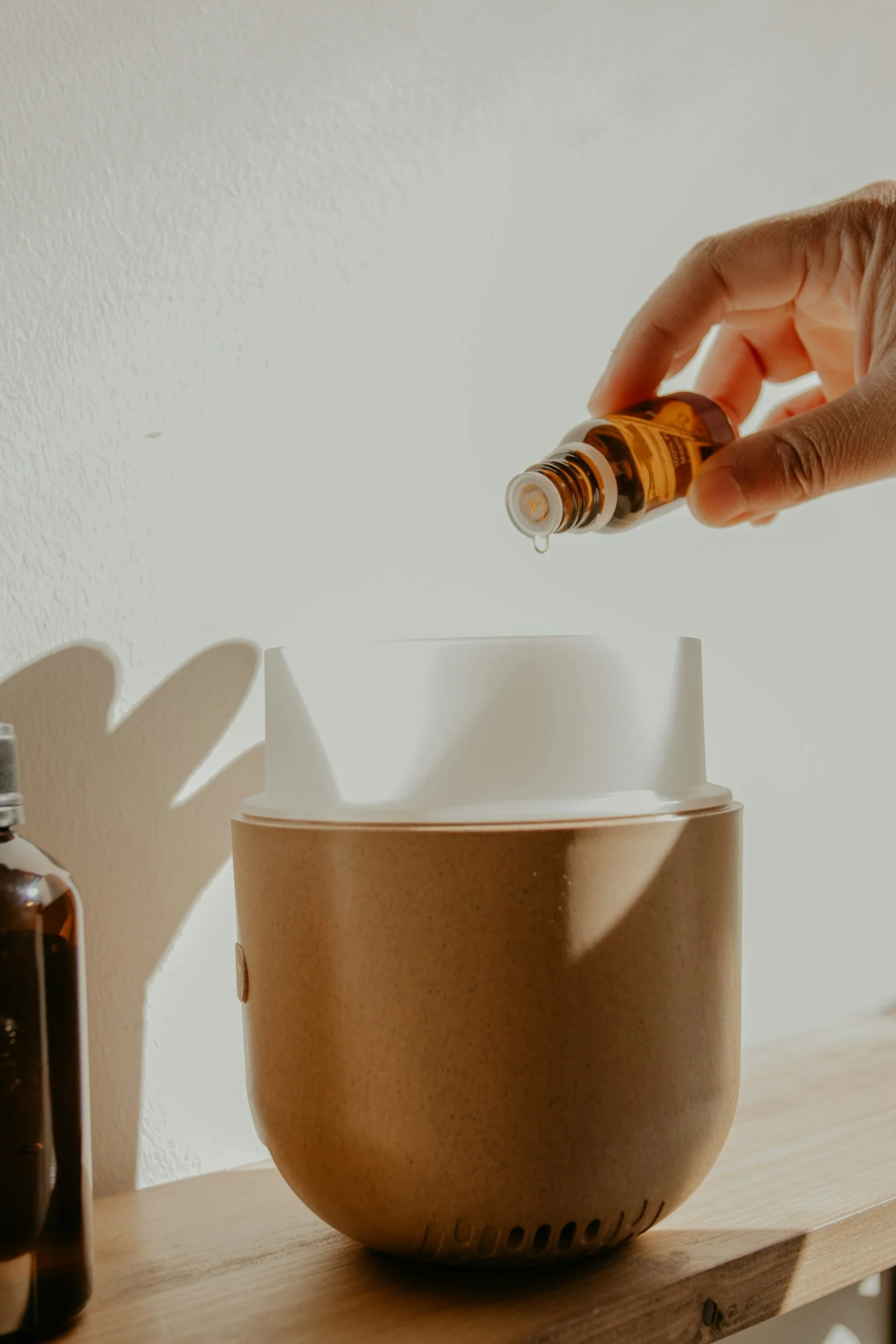 A hand is pouring a liquid from a brown essential oil bottle into a white and beige diffuser on a wooden surface, with sunlight casting shadows.