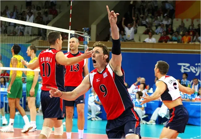 A volleyball team celebrates on the court during a match, with players wearing red and navy uniforms and a crowd watching in the background.