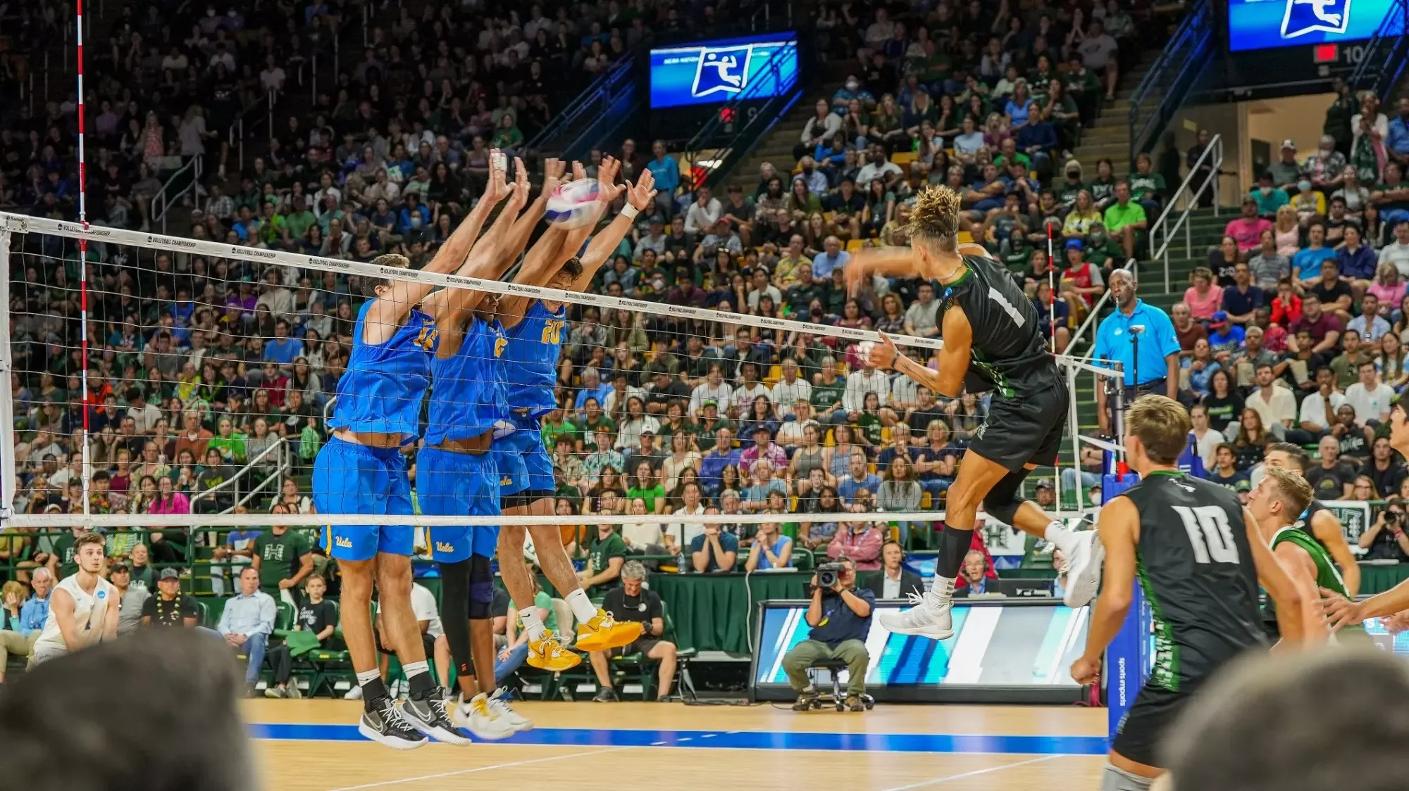 Volleyball players in action during a match; three players in blue jerseys jump to block the ball at the net while a player in black jumps to hit the ball, with a crowd watching in the background.