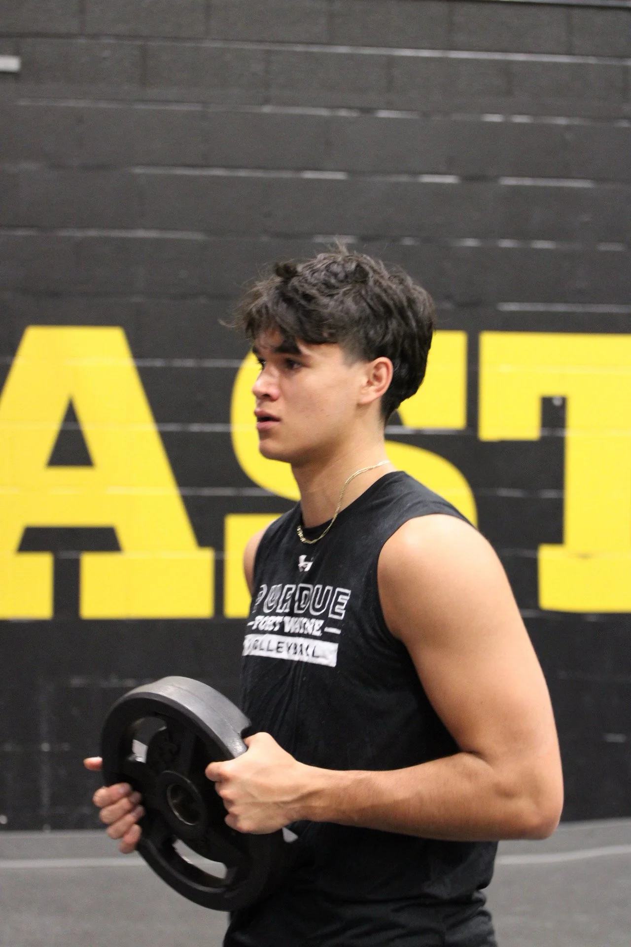 A young man at the gym holding a weight plate in his hands, wearing a black sleeveless shirt with lettering on it, and standing in front of a black and yellow wall.