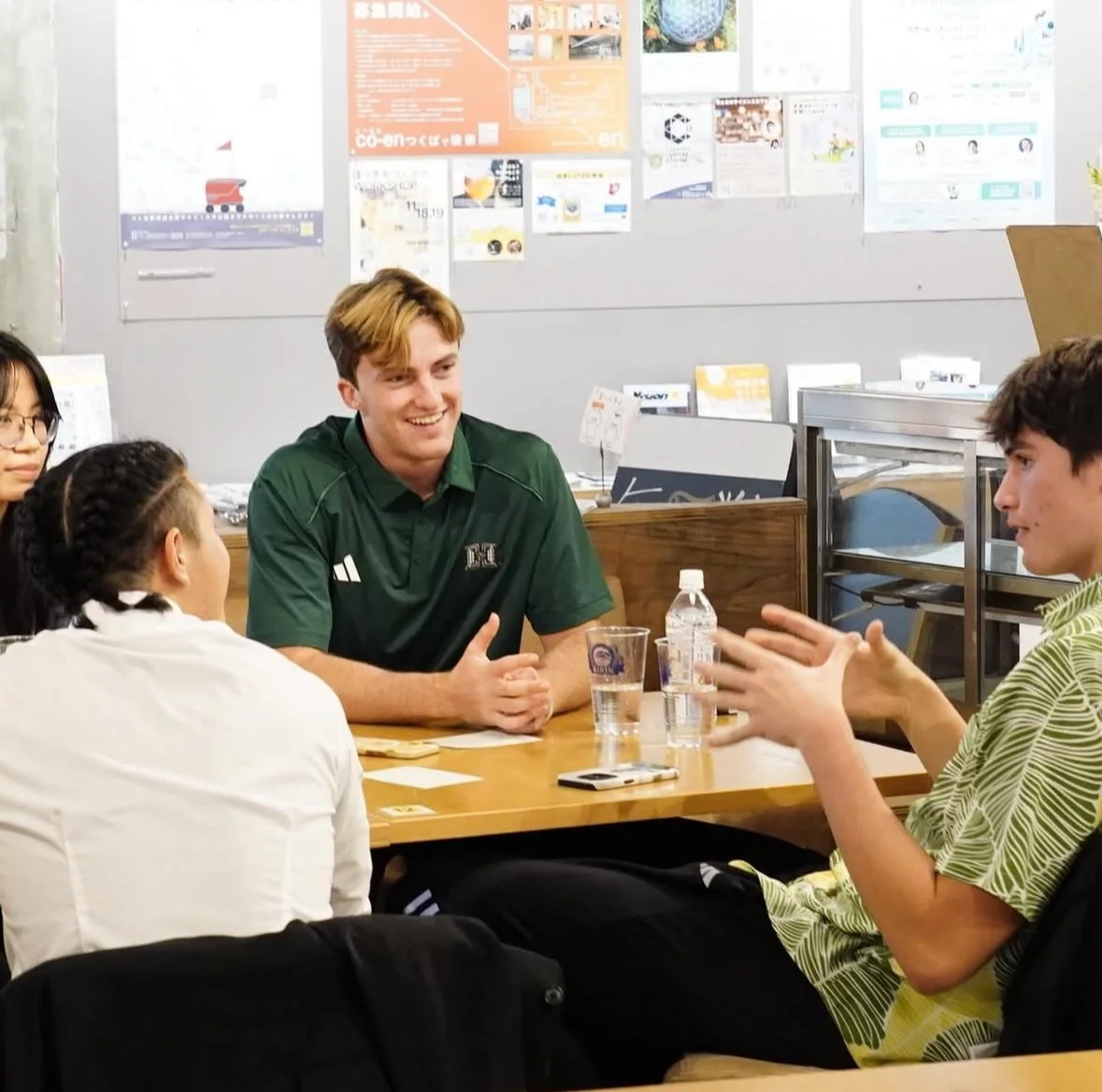 A group of people sitting around a table engaged in conversation, with a young man in a green shirt smiling and talking to a young man in a patterned yellow shirt, in a casual indoor setting with posters on the wall behind them.