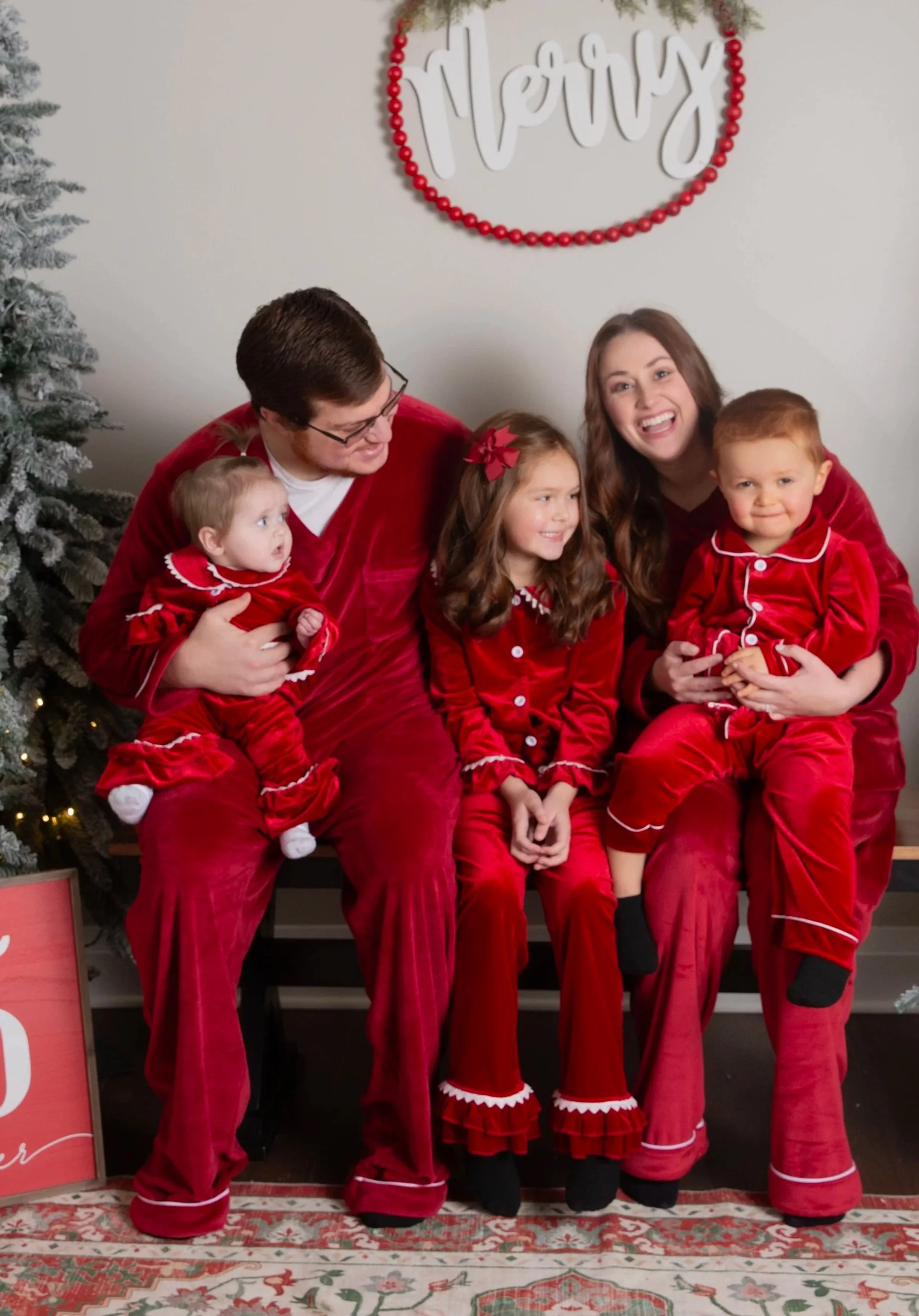 A family of six dressed in matching red pajamas posing for a Christmas photo in front of a decorated Christmas tree and a sign that says "Merry" on the wall.