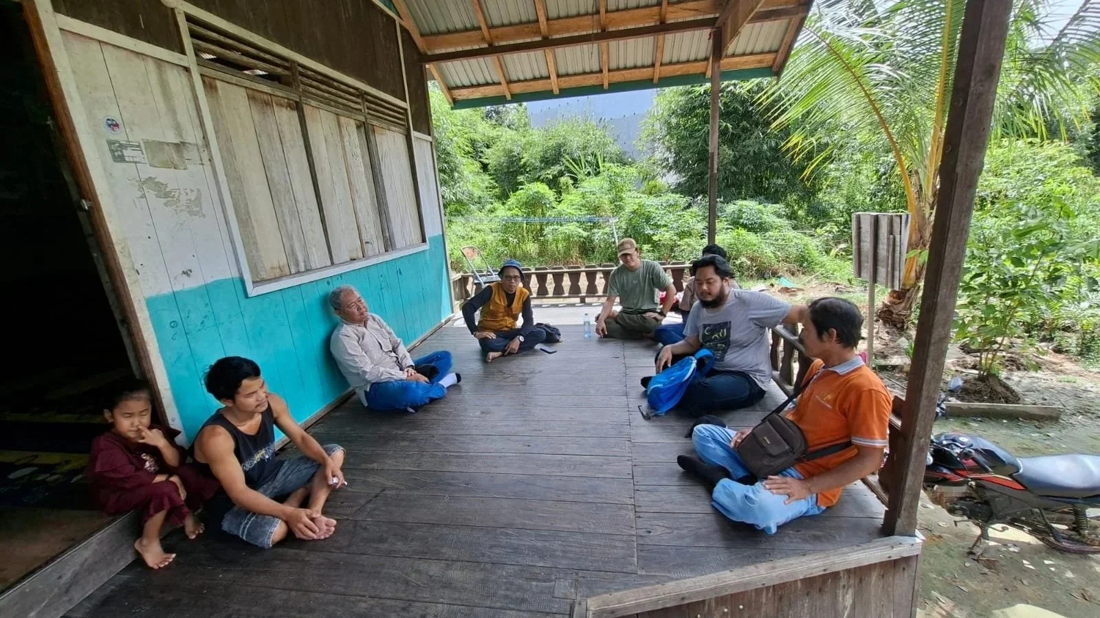 Group of nine people sitting on a wooden porch of a house, surrounded by green trees and plants, suggesting an outdoor or rural setting.