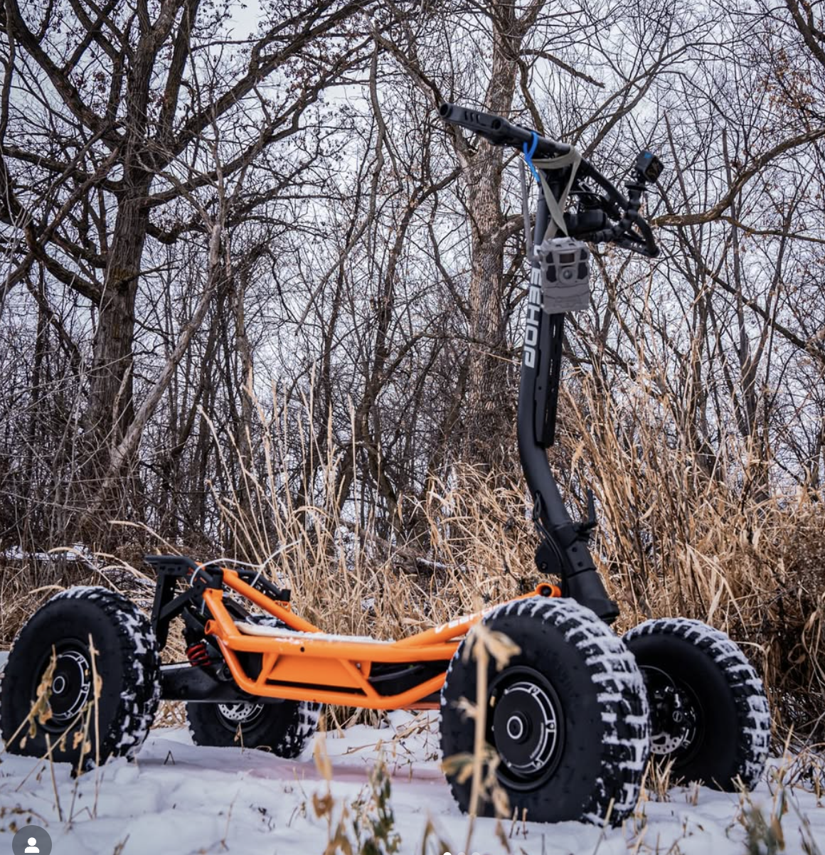 An off-road electric scooter with orange frame and large knobby tires in a snowy outdoor setting with leafless trees and dry grass.