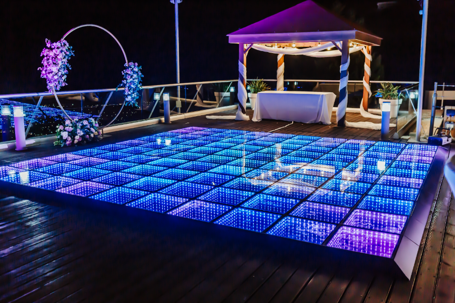Illuminated dance floor at night with blue and purple LED tiles, decorated arch with flowers, and a small pavilion with white table and plants.