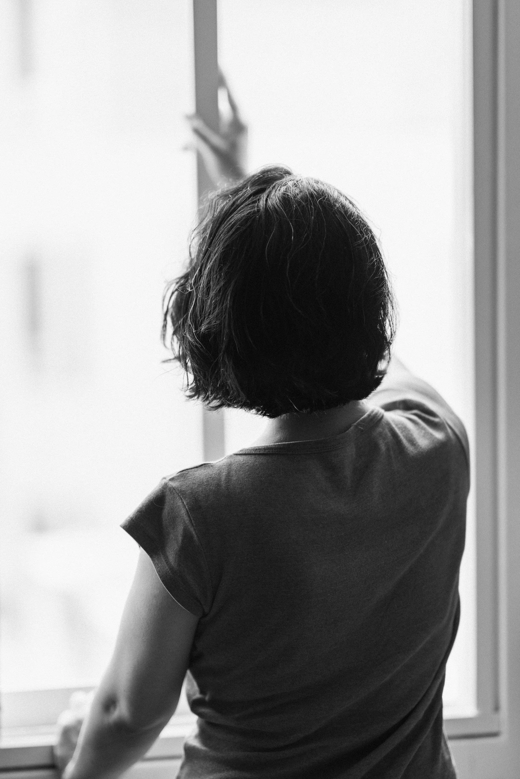 A person with short hair is reaching out toward a window or mirror with their right hand. They are wearing a short-sleeved shirt and are facing away from the camera. The setting appears to be indoors.