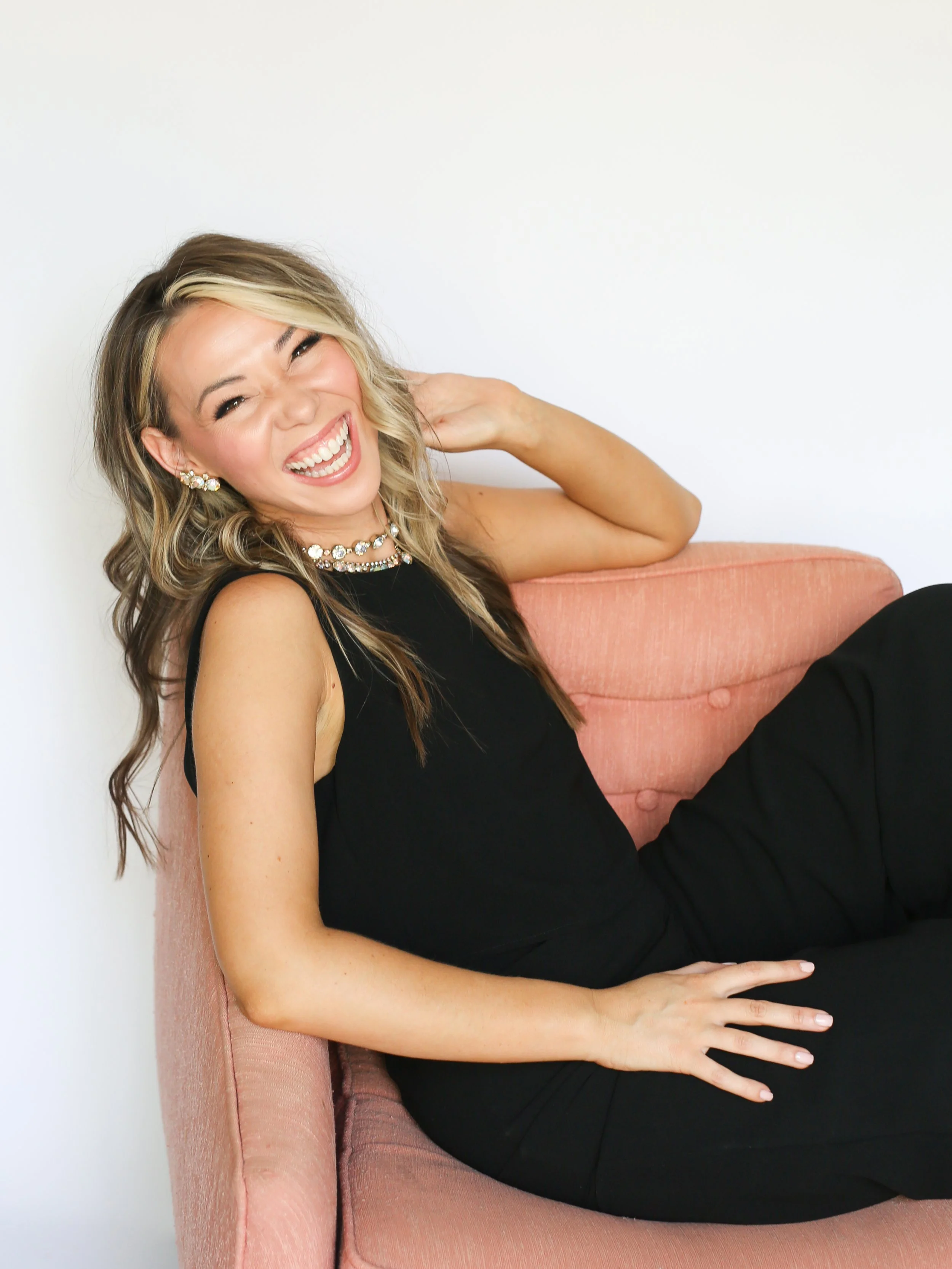 A woman with blonde hair, wearing a black dress and jewelry, sitting on a pink couch and laughing with her hand behind her neck.