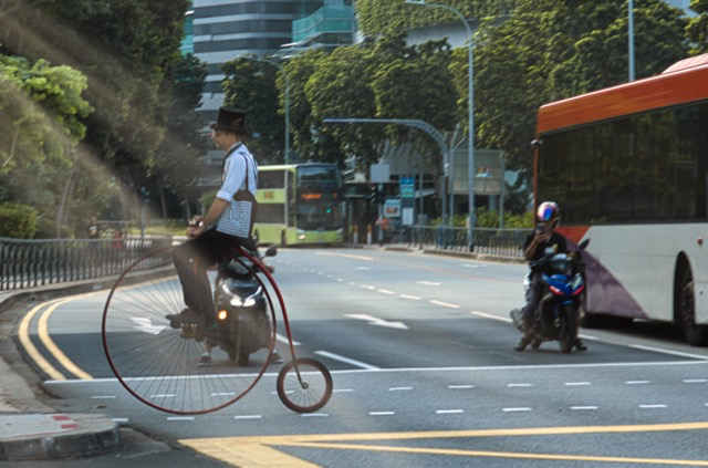 A person riding a vintage-style bicycle with a large front wheel and smaller rear wheel across an urban street, with modern buses and trees in the background.
