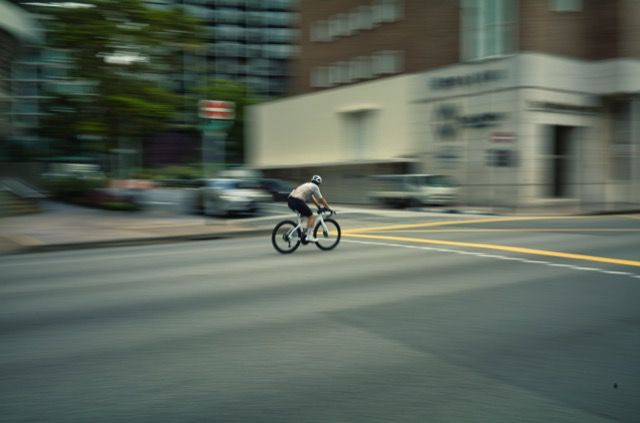 A person riding a bicycle across a city street with buildings and parked cars in the background.