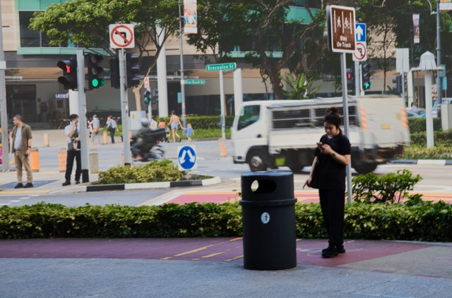 A person standing on a sidewalk next to a black trash can, using a smartphone. In the background, there is a busy street with pedestrians, cars, and motorcycles, along with traffic lights and street signs.