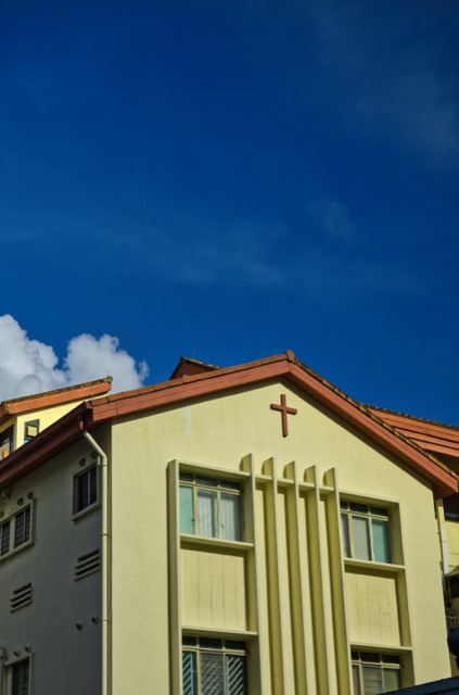 A light yellow building with a red roof and a small cross on the gable end, under a clear blue sky.
