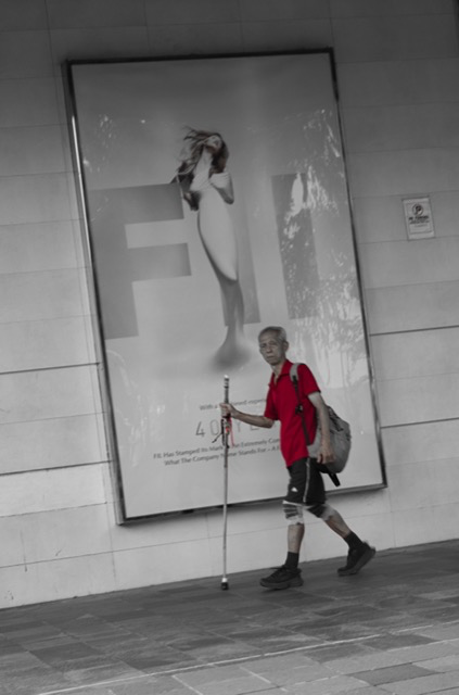 A young man with a cane and backpack walking past a large advertisement poster featuring a woman with long hair and a white dress.