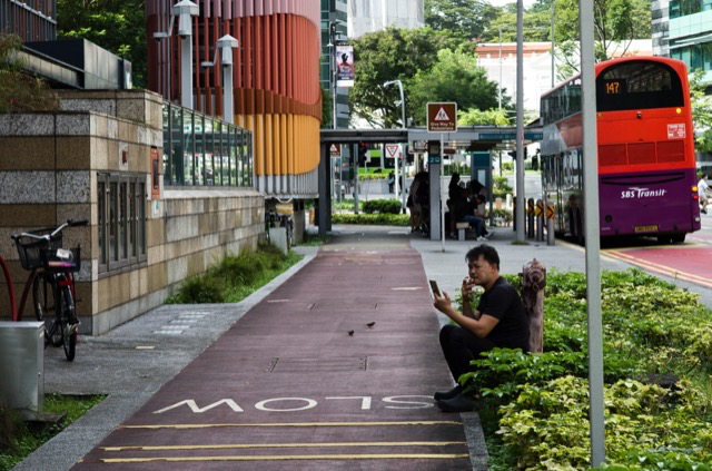 A city bus stop with people waiting, a man sitting on a bench looking at his phone, and a red and purple bus passing by.