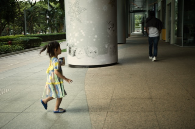 A young girl in a colorful dress and blue shoes walking on a sidewalk outside a building, holding a phone, with a man walking away inside the building.