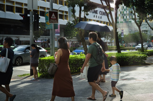 People walking on a city sidewalk in the daytime, with cars and buildings in the background, some holding umbrellas to shield from the sun.