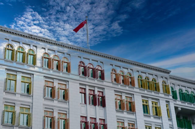 A historic white building with multiple windows, some opened, with colorful window frames in yellow, green, red, and orange, under a partly cloudy blue sky with a flag flying on top.