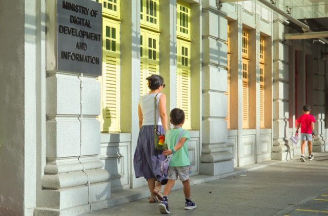 A woman and a young boy walking past a building with a sign that reads 'Ministry of Digital Development and Information.' The building has bright yellow shutters and concrete columns. Another child walking ahead is wearing a red shirt.