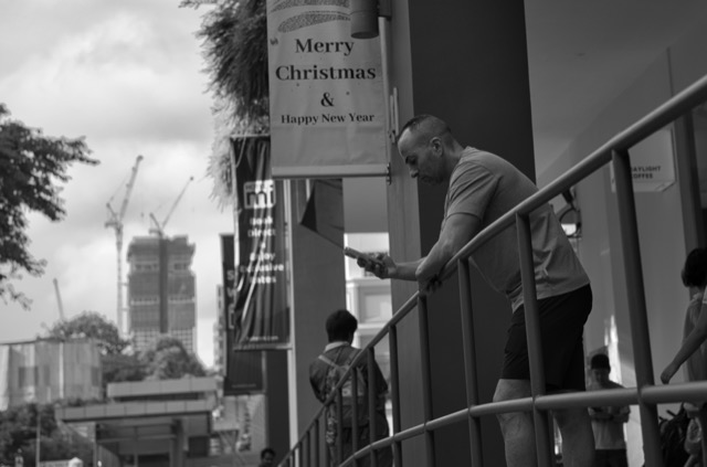 Man leaning on a railing while looking at his phone outside a building, with a Christmas and New Year banner in the background, and a cityscape with cranes and high-rise buildings.