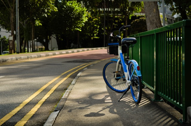 Blue bicycle locked to a green railing on a sidewalk with a street and trees in the background.