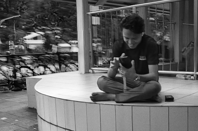 A young man sitting cross-legged on a rounded platform, looking at his phone and smiling.