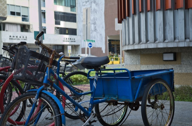 A blue cargo tricycle parked on the sidewalk with bicycles nearby in an urban area.
