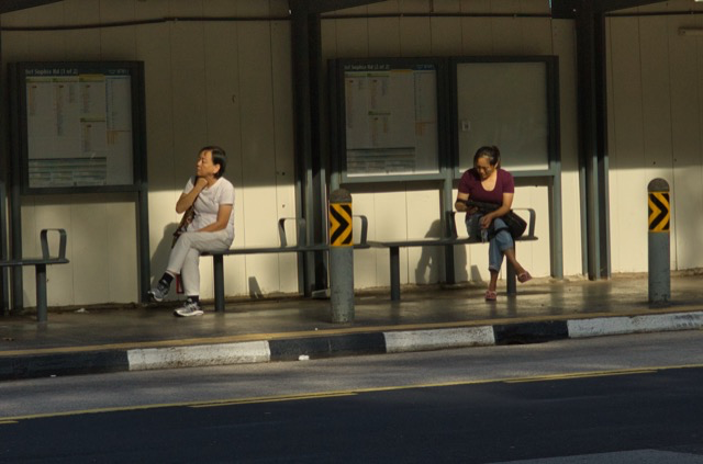 Two women sitting on separated benches at a bus stop, waiting, with bus schedule posters behind them and a pole between them on a sidewalk in sunlight.