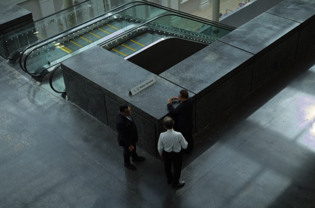 Three men are standing and talking in a modern building with glass and concrete features.
