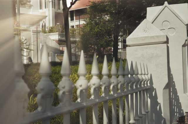 A white decorative wrought iron fence with pointed tops in front of a garden and residential buildings.
