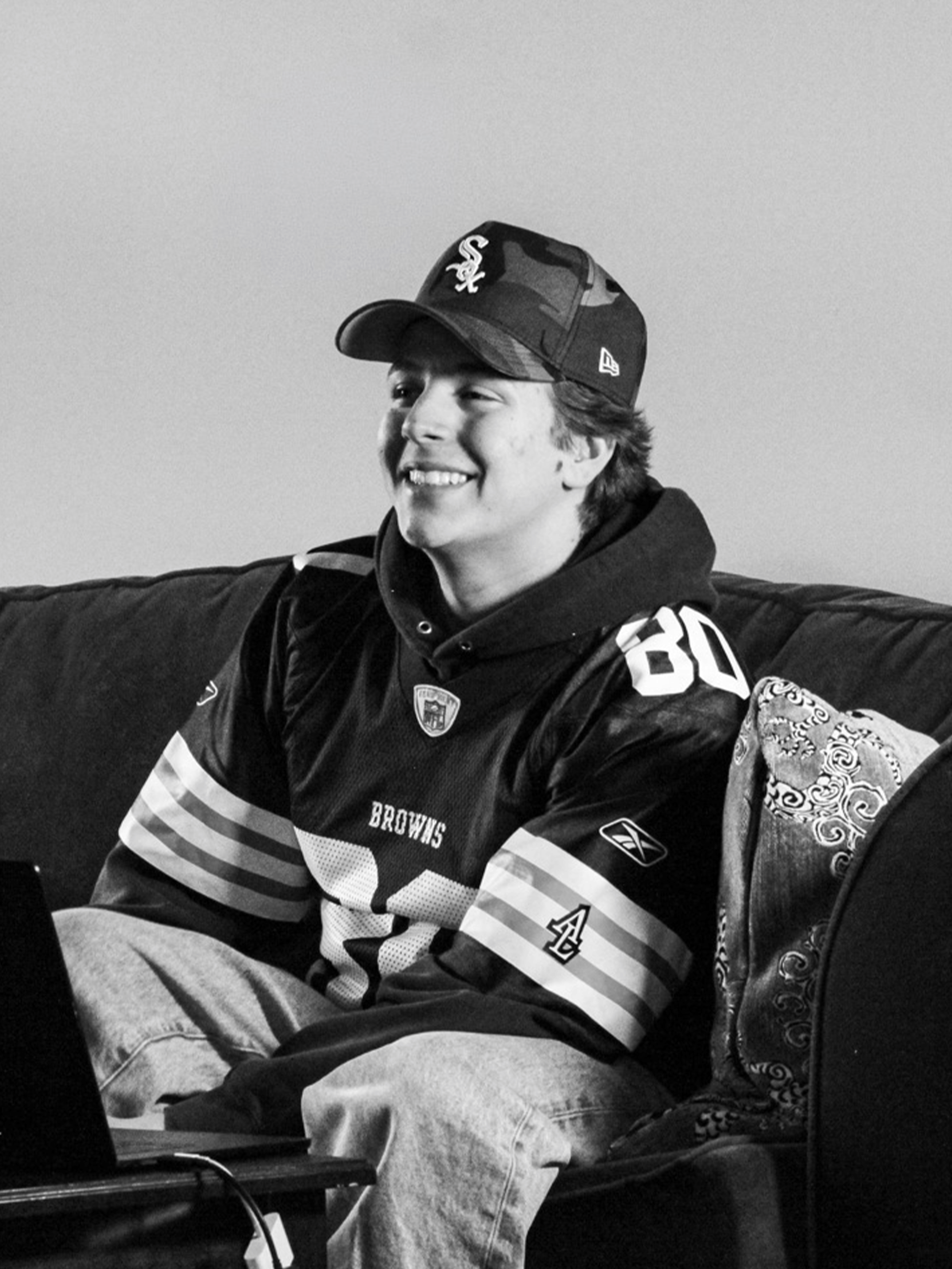 A young man wearing a football jersey, hoodie, and camouflage cap sitting on a couch and smiling, with a laptop in front of him.